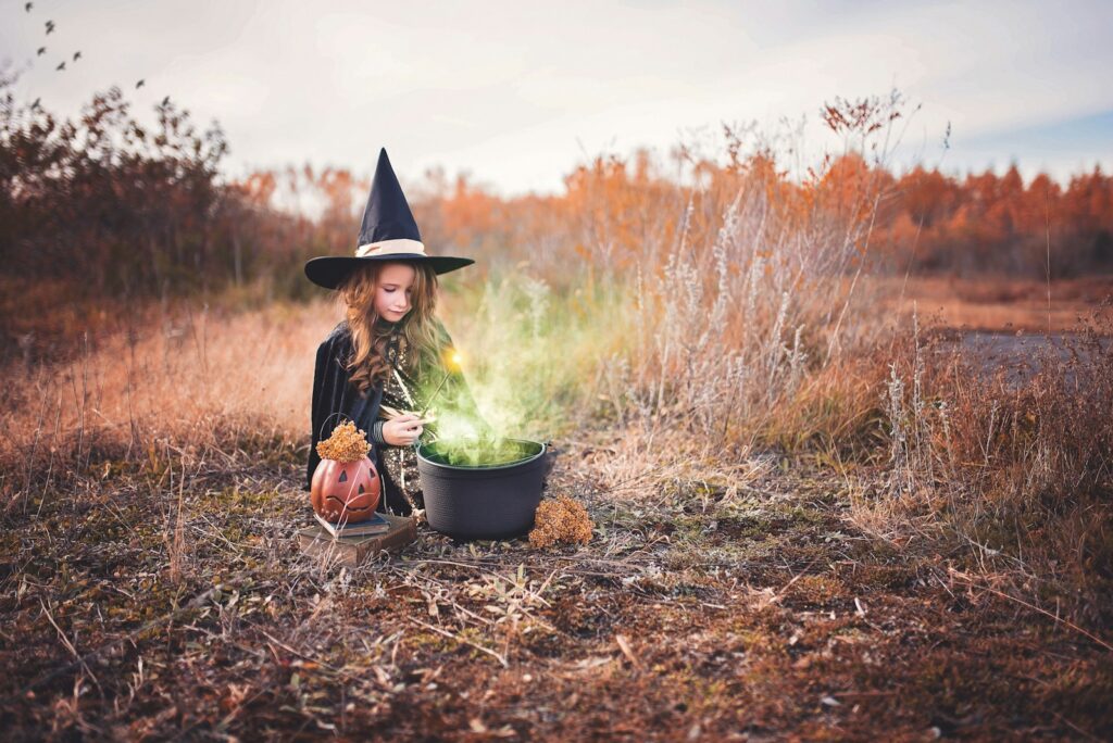 Young girl in a witch costume sitting beside a cauldron outdoors in the fall, representing a calm and gentle approach to Halloween for anxious kids.